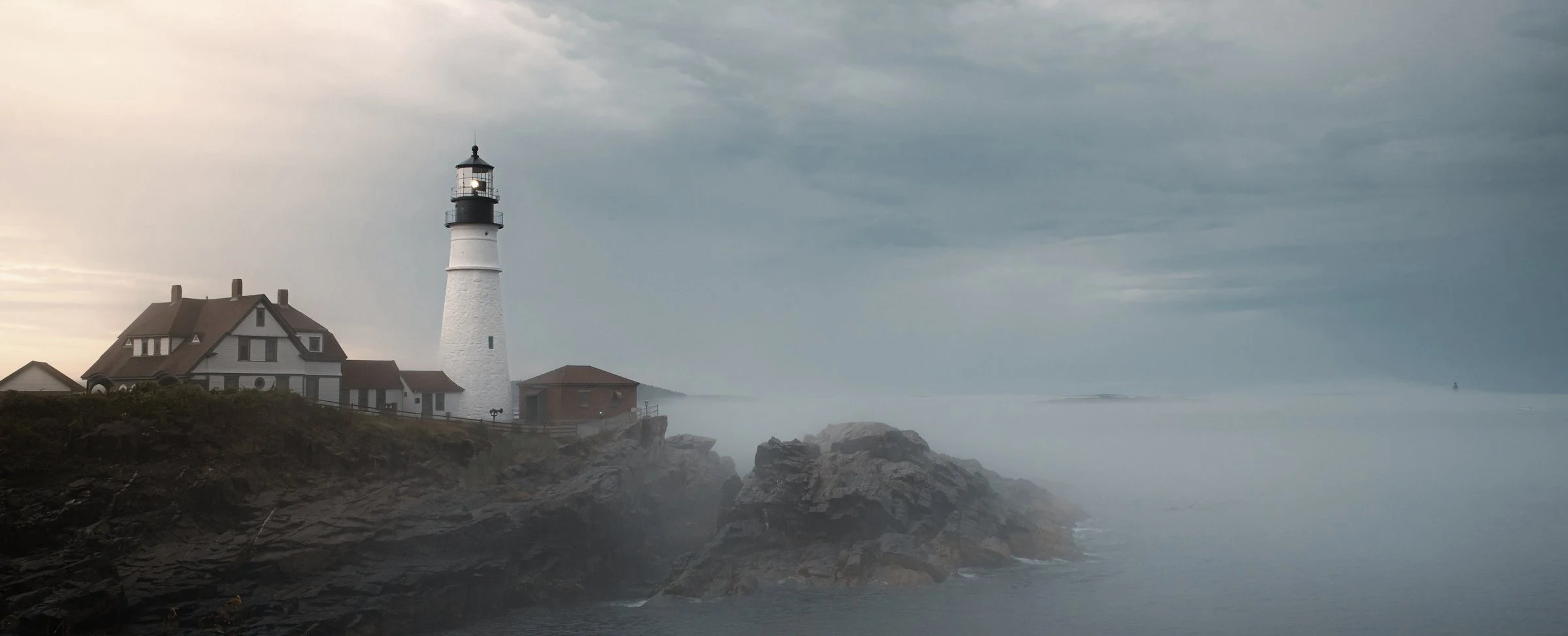 Kayalık bir burunda deniz feneri ve bekçi evi, sise gömülü — a lighthouse and its keeper’s house on a rocky headland, half-lost in sea fog